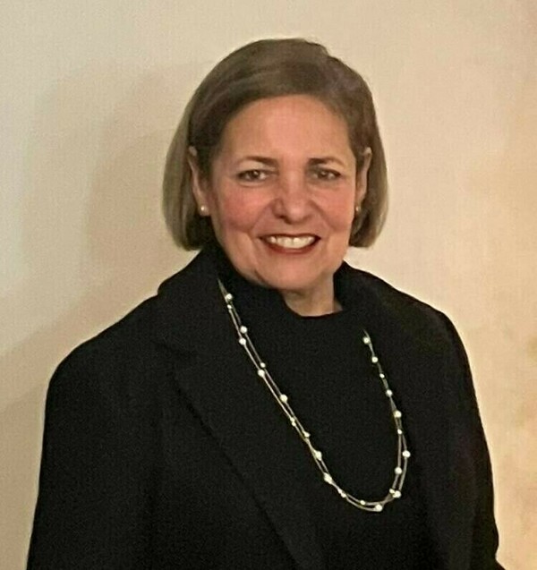 Woman with short graying hair, wearing a black turtleneck and jacket, smiles at the camera.  She wears a double-stranded pearl necklace.