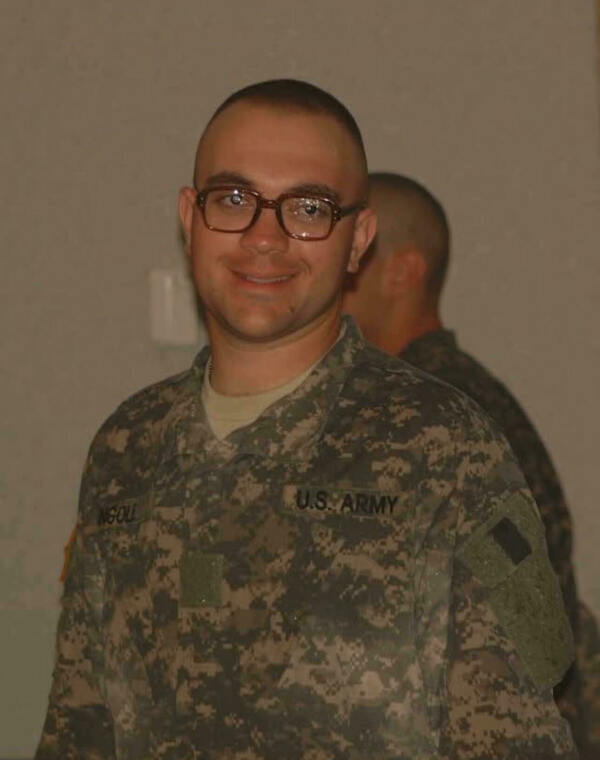 A man with a buzz cut, brown glasses, and a wide smile wears a U.S. Army camouflage uniform with "U.S. ARMY" and "NGOLL" name tapes. A blurry uniformed person stands behind him.